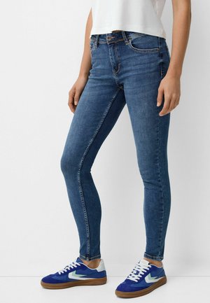 Dunne blauwe denim jeans met een medium hoge taille, voorzien van een ritssluiting en een vijf-pocket ontwerp. Gemaakt in combinatie met blauwe sportieve sneakers.