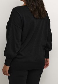 Pull noir texturé à coupe décontractée, épaules tombantes et poignets froncés. Pantalon noir partiellement visible.