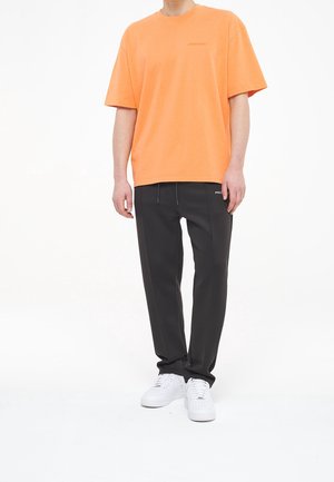 Man in een oranje T-shirt met korte mouwen, zwarte broek met trekkoord en witte sneakers, staand tegen een witte achtergrond.
