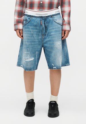 Persoană care poartă pantaloni scurți din denim uzat până la genunchi, adidași negri, șosete albe și o cămașă în carouri roșie cu branding vizibil la talie.