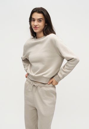 Jeune femme aux longs cheveux bruns portant un sweat-shirt beige clair et un pantalon de survêtement assorti, debout les mains sur les hanches.
