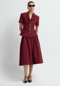 Blazer bordeaux ajusté à manches courtes, boutons dorés et taille peplum, associé à une jupe midi assortie ; chaussures noires à bout pointu.