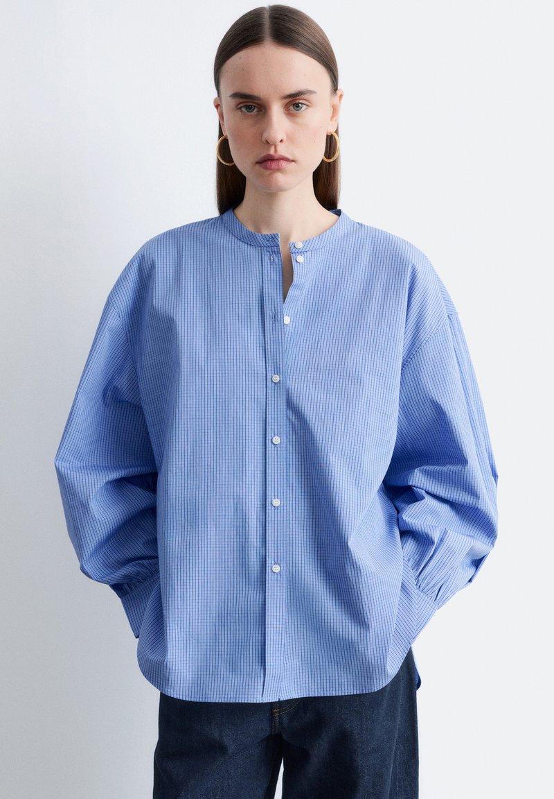 Femme aux cheveux bruns raides portant une chemise à carreaux bleue claire ample à boutons avec col montant et boucles d'oreilles en forme d'anneaux dorés, sur fond blanc.