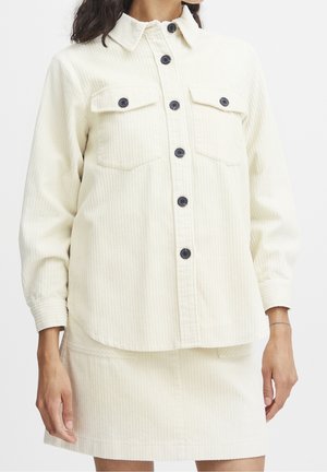 Kobieta ubrana w kremową sztruksową koszulę zapinaną na guziki z przednimi kieszeniami oraz dopasowaną mini spódnicę na jednolitym tle.