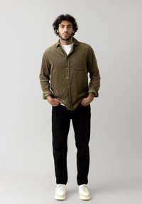 Oliven grøn corduroy skjorte med en forlomma, parret med sorte jeans og hvide sneakers. Modellen står med hænderne i lommerne foran en neutral baggrund.