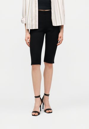 Person iført sorte, knælange tætsiddende shorts, sorte højhælede sandaler med ankelremme og en hvid stribet jakke.