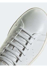Vit lädersneaker med rund tå, ton-i-ton snörning, perforerade detaljer och en ljusfärgad gummisula. Slitstark yta, minimalistisk design.