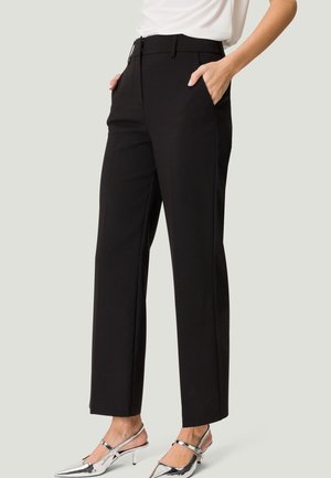 Mujer con pantalones negros de pierna recta, con las manos en los bolsillos y tacones plateados de punta fina sobre un fondo liso.