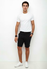 White crew-neck t-shirt paired with dark shorts featuring a button closure and side pockets. Worn with white sneakers, standing against a white background.