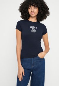 Tumesinine soonikkoes lühikeste varrukatega t-särk, millel on valge tekst: "TOMMY 85 AUTHENTIC ORIGINAL." Lähestikku istuv lõige ja ümara kaelusega.