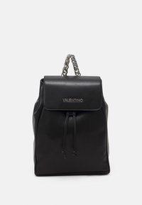 Svart läderryggsäck med klaffstängning, silverkedjehandtag, dragsko-detalj och präglad logotyp på framsidan. Slät textur.