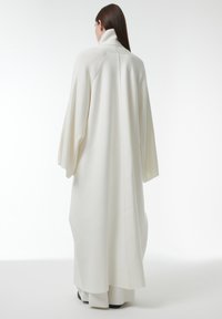 Overmaat witte gebreide jas met een hoge kraag, doorgezakte schouders en een naadloos ontwerp aan de achterkant, met een gladde textuur en mouwen van volledige lengte.
