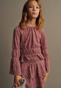 Vestito a maniche lunghe, con vita smock in rosa profondo e stampa floreale. Polsini e vita con volant; la modella tiene un accessorio grigio lavorato a maglia.