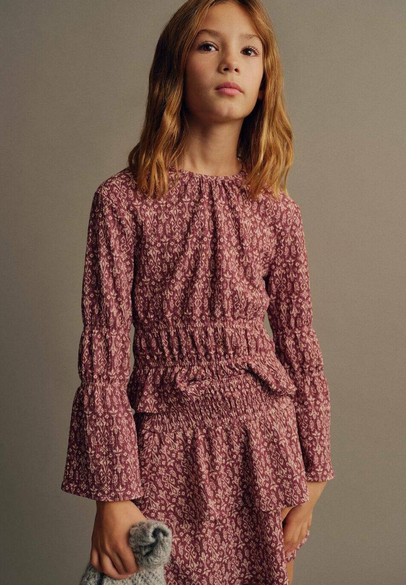 Vestito a maniche lunghe, con vita smock in rosa profondo e stampa floreale. Polsini e vita con volant; la modella tiene un accessorio grigio lavorato a maglia.