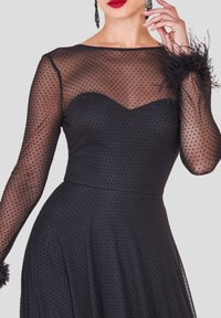 Musta mekko, jossa on läpikuultava pilkullinen hihamateriaali, sydämenmuotoinen pääntie, sulkaiset koristeet kalvosimissa, vartalonmyötäinen yläosa ja liehukehelmainen hame.