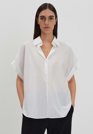 Chemise blanche à manches courtes avec un col, présentant un décolleté en V et une coupe ample. Texture lisse avec des poignets retroussés et un minimum de détails.