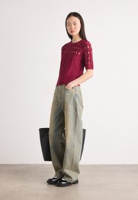 Cremfarbener gestrickter Oberteil mit Ausschnitten, kombiniert mit lockeren, ausgebleichten blauen Jeans. Eine schwarze Tragetasche und klobige schwarze Schuhe runden den Look ab.
