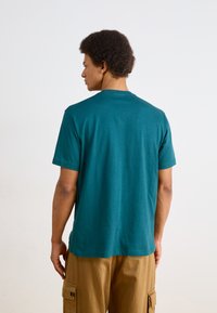 T-shirt de algodão teal com mangas curtas, gola redonda e corte reto. Design simples, textura suave, com um pequeno detalhe perto da gola.