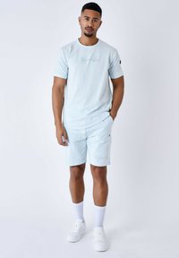 T-shirt di cotone azzurro chiaro con logo frontale, abbinato a pantaloni corti coordinati e sneaker bianche. Il modello è in piedi su uno sfondo neutro.