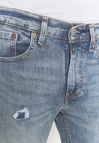 Primer plano de unos vaqueros de mezclilla azul de corte medio con un pequeño detalle rasgado en el muslo izquierdo y botones y lazos del cinturón visibles.