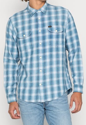Homme portant une chemise à carreaux bleu clair et blanc à manches longues avec deux poches à rabat à l'avant et un jean bleu clair.
