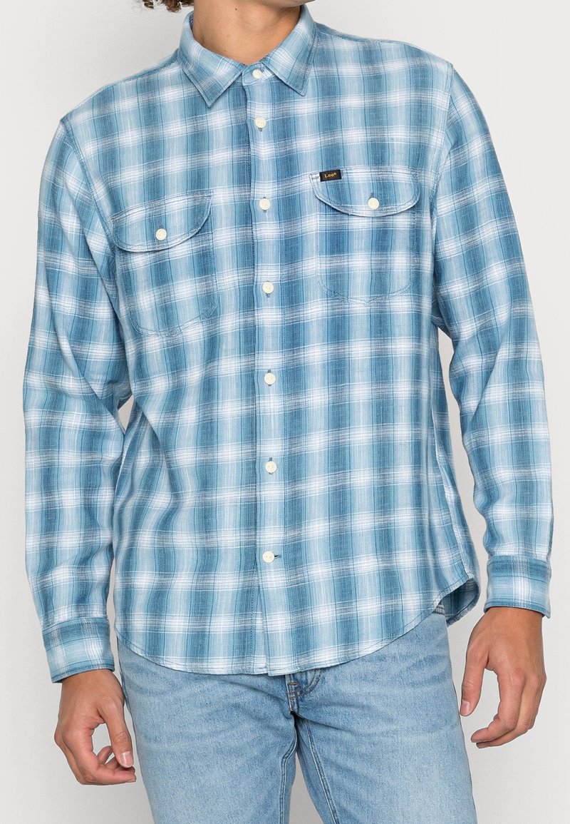 Homme portant une chemise à carreaux bleu clair et blanc à manches longues avec deux poches à rabat à l'avant et un jean bleu clair.