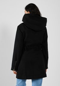 Manteau noir à capuche avec une taille ceinturée, des zips sur les côtés, une coupe adaptée et une texture lisse. Porté par-dessus un jean large bleu clair.