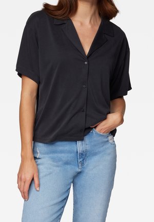 Kvinde iført sort kortærmet skjorte med krave og lyseblå revnede jeans, den ene hånd i lommen og den anden hængende ned.
