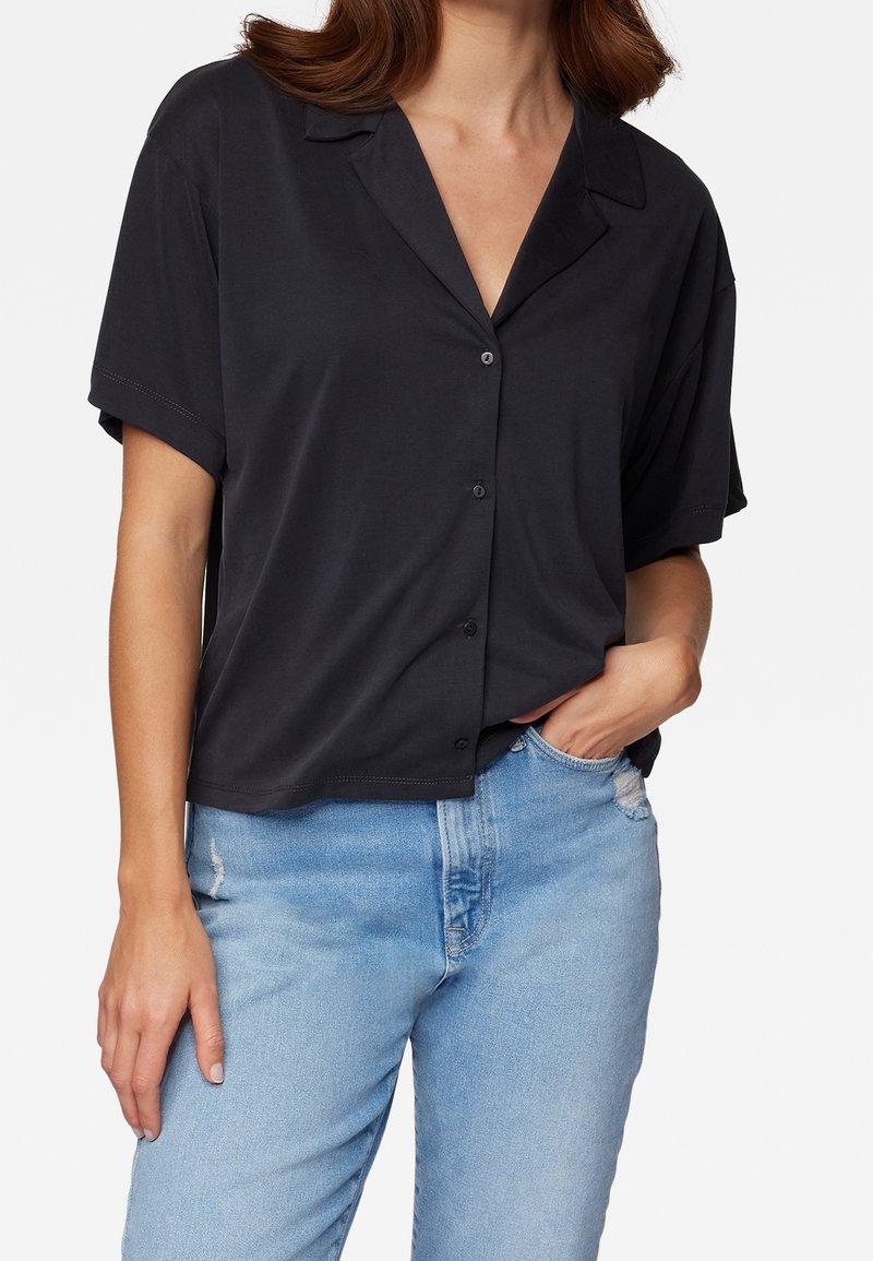 Femme portant une chemise noire à manches courtes avec col et un jean bleu clair déchiré, une main dans la poche et l'autre vers le bas.