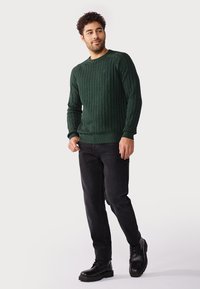 Pull en maille torsadée verte avec un col et des poignets côtelés, porté par-dessus un jean foncé et des bottes àlacets noires, présenté sur fond blanc.