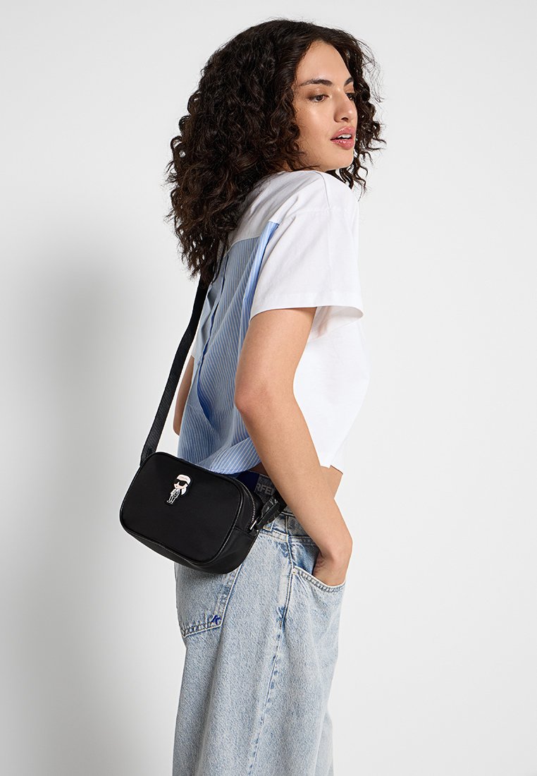 Bolso cruzado negro con cierre de cremallera, que presenta un detalle gráfico de dibujos animados y un acabado texturizado. Lleva puesto un modelo con un atuendo casual.