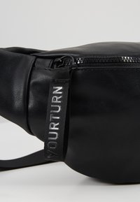 Sac en cuir noir avec une fermeture éclair et une languette noire sur laquelle est inscrit le texte « YOURTURN » en lettres blanches.