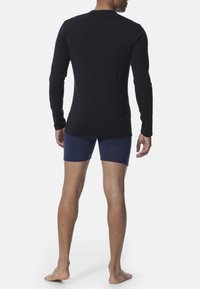Long-sleeve black t-shirt made of smooth fabric, paired with navy blue boxer shorts. Model stands barefoot, showing a slim profile from behind.