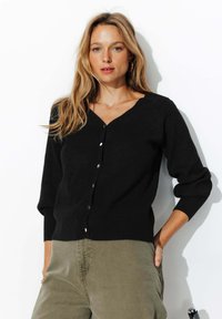 Cardigan noir en tricot avec un col en V et des boutons dorés, doté de manches longues. Porté par-dessus un pantalon olive, sur fond blanc.