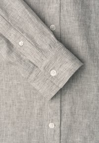Chemise en tissu gris clair texturé avec des boutons blancs, montrant une manche pliée et des détails de poignets boutonnés.