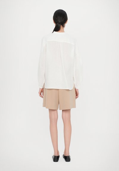 Hvit bluse med lange ermer og avslappet passform, kombinert med beige skreddersydde shorts og svarte slip-on-sko. Enkel og lett design.