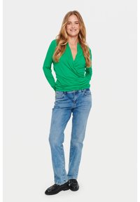 Top avvolgente verde a maniche lunghe con dettaglio drappeggiato frontale, abbinato a jeans a gamba dritta azzurri chiari e scarpe nere. Sfondo bianco.