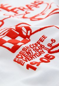 Tissu blanc avec texte rouge brodé et graphismes, comprenant "FRESH COFFEE EVENT 19-05 EVERYDAY 9-5" et un design stylisé de tasse de café.