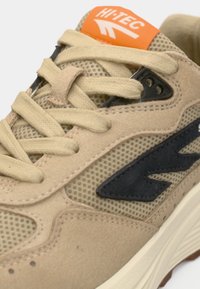 Μπεζ αθλητικό αθλητικό παπούτσι με υλικά από δίχτυ και σουέντ, μαύρα διακοσμητικά, μπεζ κορδόνια και πορτοκαλί λογότυπο στη γλώσσα.