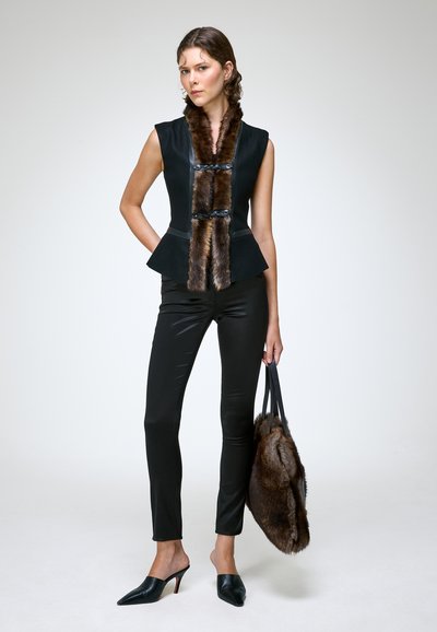 Femme portant un top noir sans manches avec une bordure en fourrure marron, pantalon noir, talons pointus noirs, et tenant un grand sac à main en fourrure marron.