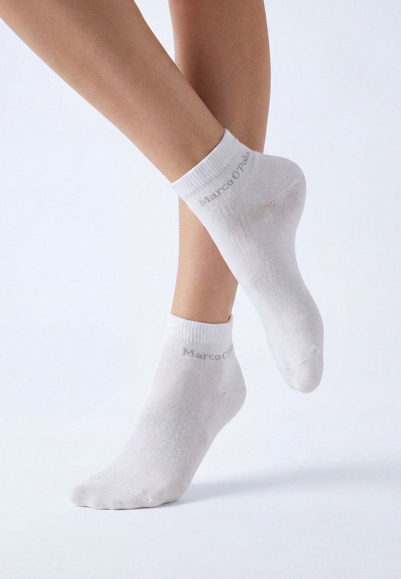 Pieds portant des chaussettes blanches à la cheville avec le texte « Marco O'Polo », posés sur un fond clair uni avec un pied légèrement levé.