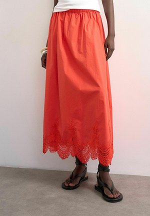 Personne portant une jupe midi orange vif avec un ourlet en dentelle, un haut blanc, des sandales noires à brides, et des bracelets au poignet gauche, debout à l'intérieur.