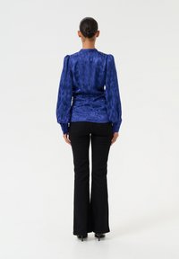 Blue satin blouse with textured finish, puffed sleeves, cinched waist, paired with black flared trousers and high-heeled shoes.