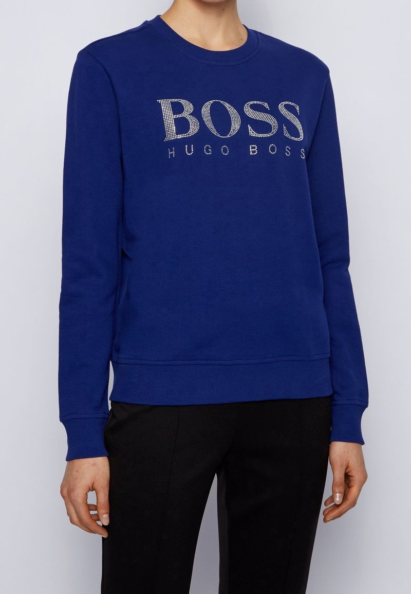 Personne portant un sweat-shirt bleu à manches longues avec « BOSS HUGO BOSS » en strass sur la poitrine et un pantalon noir.