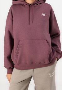 Sudadera con capucha púrpura y bolsillo estilo canguro con puños acanalados. Presenta un logo blanco "NB" en el pecho. Combinada con pantalones de chándal gris claro.