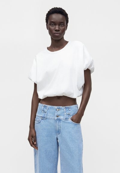 Femme portant un t-shirt blanc ample et court ainsi qu'un jean bleu clair taille haute, la main dans la poche, debout devant un fond uni.