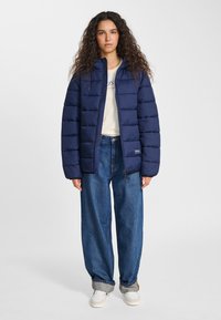 Marinblå vadderad jacka med quiltad design, dragkedja och elastiska muddar, bärs över en krämfärgad skjorta och kombineras med lösa blå denimbyxor.