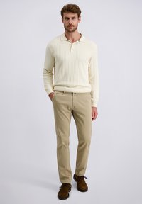 Mann steht in einem cremefarbigen, gerippten Langarm-Polo, beigefarbenen Hosen und braunen Schnürschuhen, mit Händen in den Taschen, vor einem schlichten hellen Hintergrund.