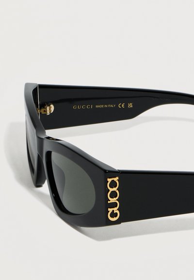 Μαύρα ορθογώνια γυαλιά ηλίου με σκούρους φακούς, με χρυσό λογότυπο Gucci στο πλαϊνό και την επιγραφή «Made in Italy» στο εσωτερικό του βραχίονα.