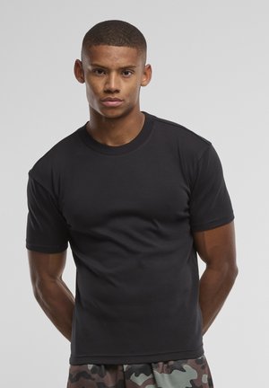 Jeune homme aux cheveux courts portant un t-shirt noir uni et un pantalon camouflage, debout devant un fond gris uni.